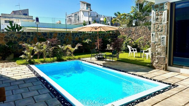 Uma piscina retangular com borda de pedra, rodeada por um terraço com cadeiras e um guarda-sol.