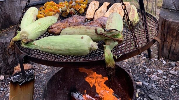 Una parrilla al aire libre con mazorcas de maíz y verduras, rodeada de árboles y una mesa de picnic en un entorno boscoso.