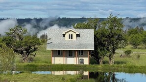 Ein eingeschossiges Haus mit Satteldach und einer Veranda. Vor der Haustür befindet sich eine kleine Veranda mit einem Stuhl. Das Haus wird von einer Wiese und einem sich spiegelnden Gewässer umgeben.