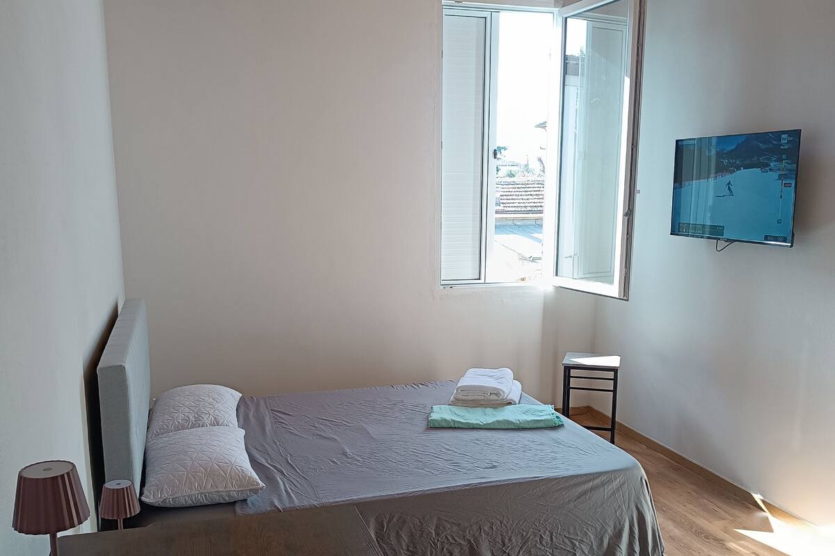 Ein Schlafzimmer mit Holzbalkendecke, einem Bett mit grauer Bettdecke, einem kleinen Nachttisch, einer Lampe und einem an der Wand befestigten Fernseher.