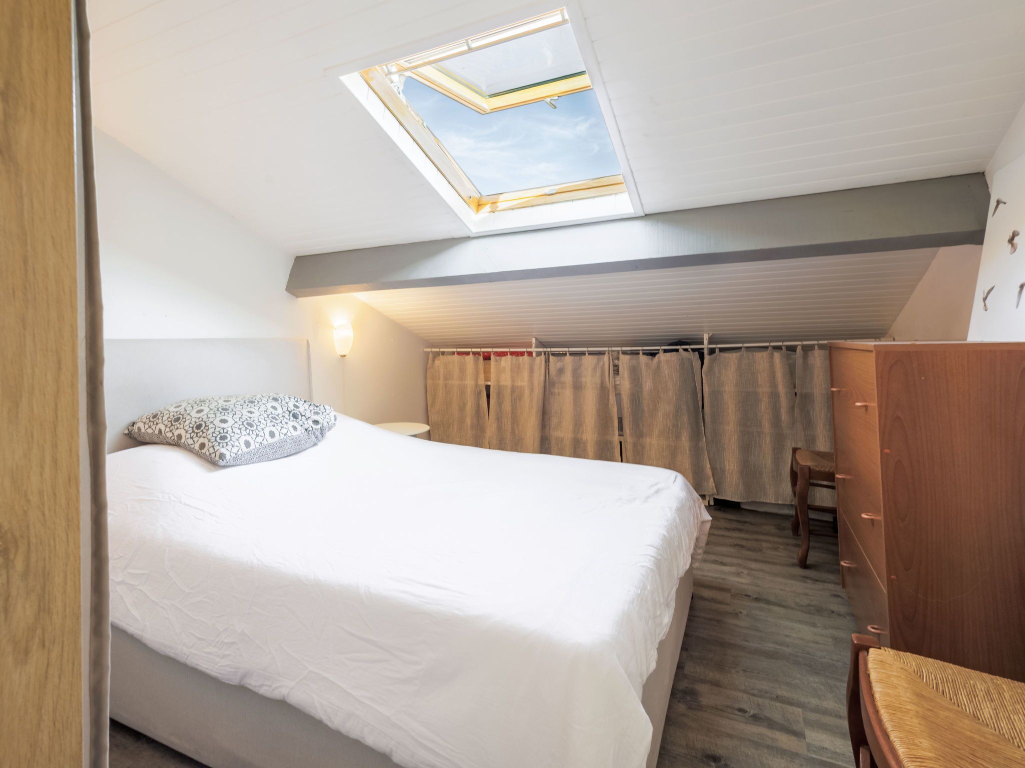 Une chambre avec un puits de lumière, un lit agrémenté d’un coussin à motifs, une commode en bois et une chaise.
