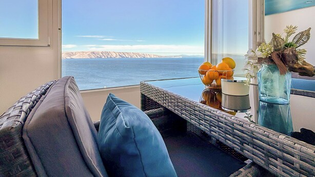 Un balcón con vistas al mar, una silla de mimbre, un cojín y un florero con flores.