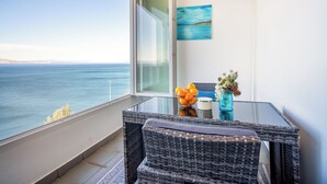 Un balcón con vista al mar, una mesa de mimbre con un florero y naranjas, y un cuadro en la pared.