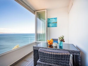 Ein Balkon mit Blick aufs Meer, ein Korbstuhl mit einer Vase und Orangen sowie ein Gemälde an der Wand.