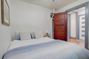 Un dormitorio con cama, mesita de noche, puerta de madera y un acceso abierto a otra habitación.