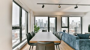 Uma sala de jantar moderna com uma mesa comprida, cadeiras pretas e um sofá azul. Grandes janelas oferecem vista para a cidade.