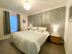 A bedroom with a large bed, a nightstand with a lamp, a window with curtains, and a floral wallpaper.