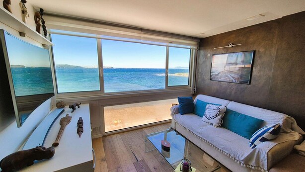 Ein Zimmer mit großem Fenster mit Blick auf das Meer, einer Couch mit blauen Kissen, einem kleinen Couchtisch und einem Holzboden.
