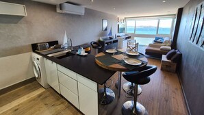 Eine moderne Küche mit einer Kochinsel, ein Essbereich mit Tisch für zwei Personen und ein Wohnbereich mit Sofa und Meerblick.