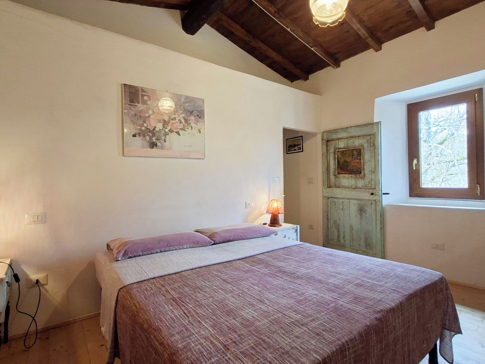 A bedroom with a bed, a lamp, a painting, a door, and a window.