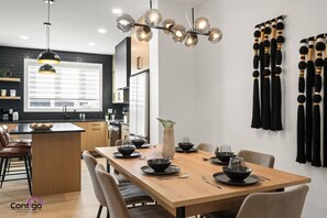 Una cocina moderna con una isla central, iluminación colgante y una zona de comedor con una mesa de madera preparada para cuatro comensales.