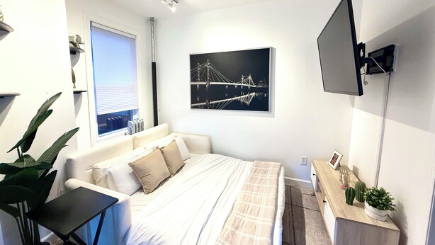 Ein modernes Schlafzimmer mit Bett, Nachttisch, Fernseher und einem Fenster mit Jalousien.