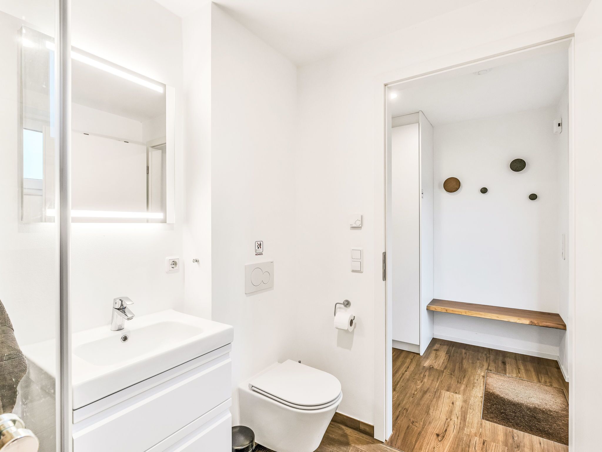 A modern bathroom with a white sink, toilet, and mirror. There is a wooden floor and a small shelf with decorative items.