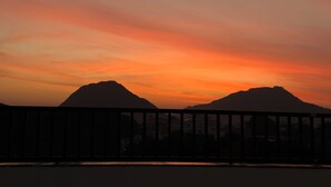 Zonsondergang met twee bergtoppen die donker afsteken tegen een kleurrijke lucht, gezien vanaf een balkon met een hekwerk.