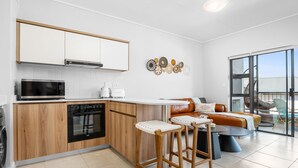 A modern kitchen with a microwave, oven, and coffee maker, a bar stool area, and a sofa with a view to the outside. - Ballito Hills 887 (Ballito)