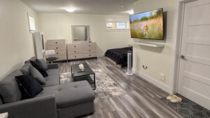 A modern living room with a grey sectional sofa, a flat-screen TV mounted on the wall, and a wooden floor. - Cozy Private Basement Suite (Halifax)