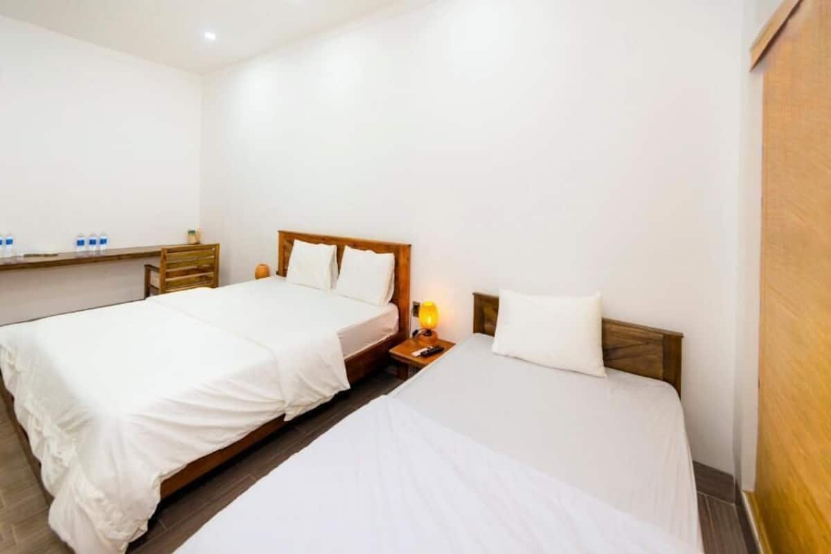 A hotel room with two single beds, wooden headboards, white bedding, a nightstand with a lamp, and a wooden shelf with water bottles.