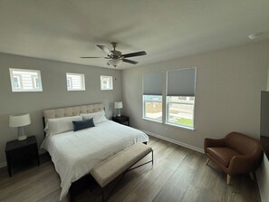 A bedroom with a bed, bedside tables, a ceiling fan, windows, and a chair. - The Hofstaat Haus: Modern NB Retreat (New Braunfels)