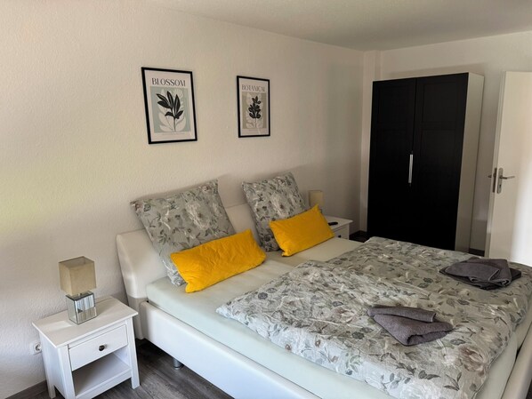 A bedroom with a bed, two pillows, a nightstand, a lamp, and two framed botanical prints on the wall. - FeWo Harzzeit - Stylish time-out in Vienenburg with terrace, WLAN & parking lot (Goslar)