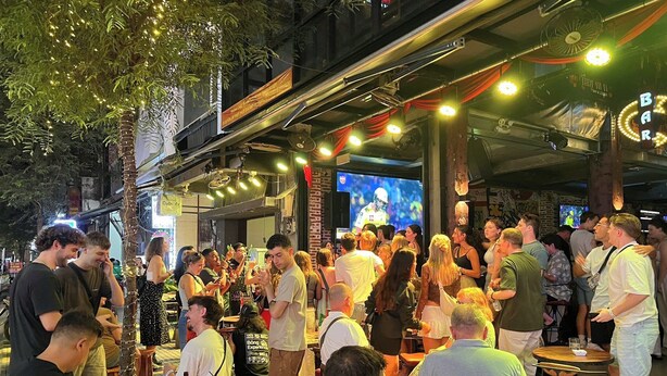 Area esterna di un bar affollato, con tavoli, sedie e un grande schermo che trasmette un evento sportivo, oltre a un cartello con il nome del bar.