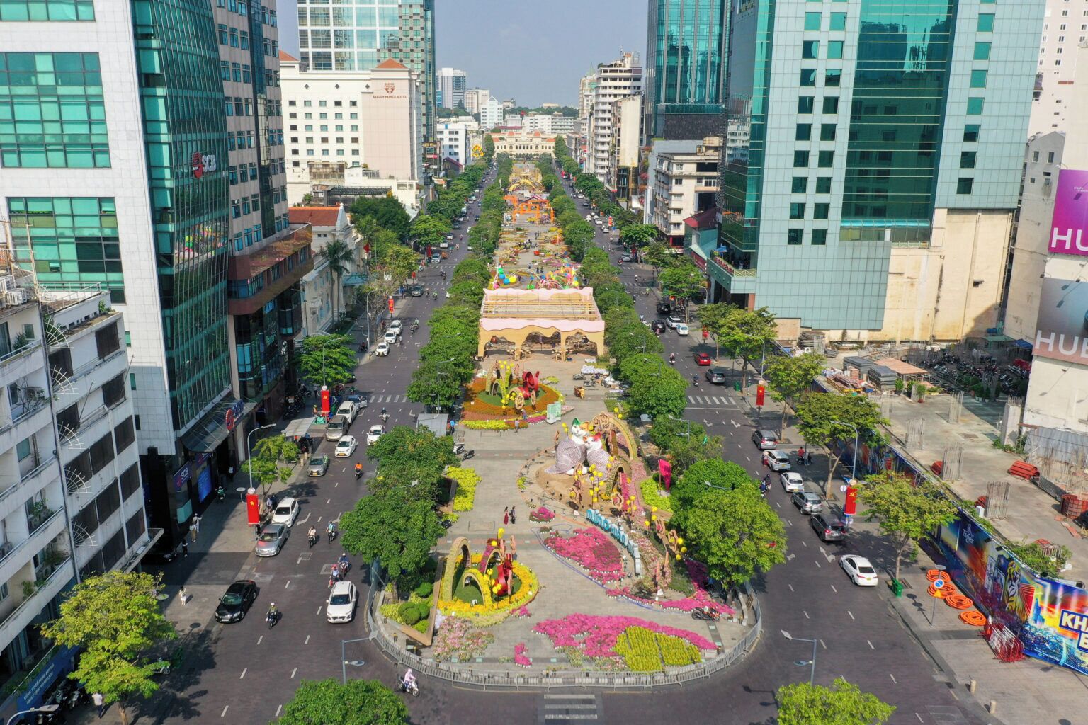 A city street with modern buildings, a central roundabout, and a large flower display.