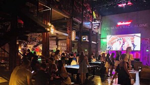 Zona bar con tavoli da biliardo e persone che giocano a biliardo. Sono presenti insegne al neon e un grande schermo che mostra uno spettacolo.