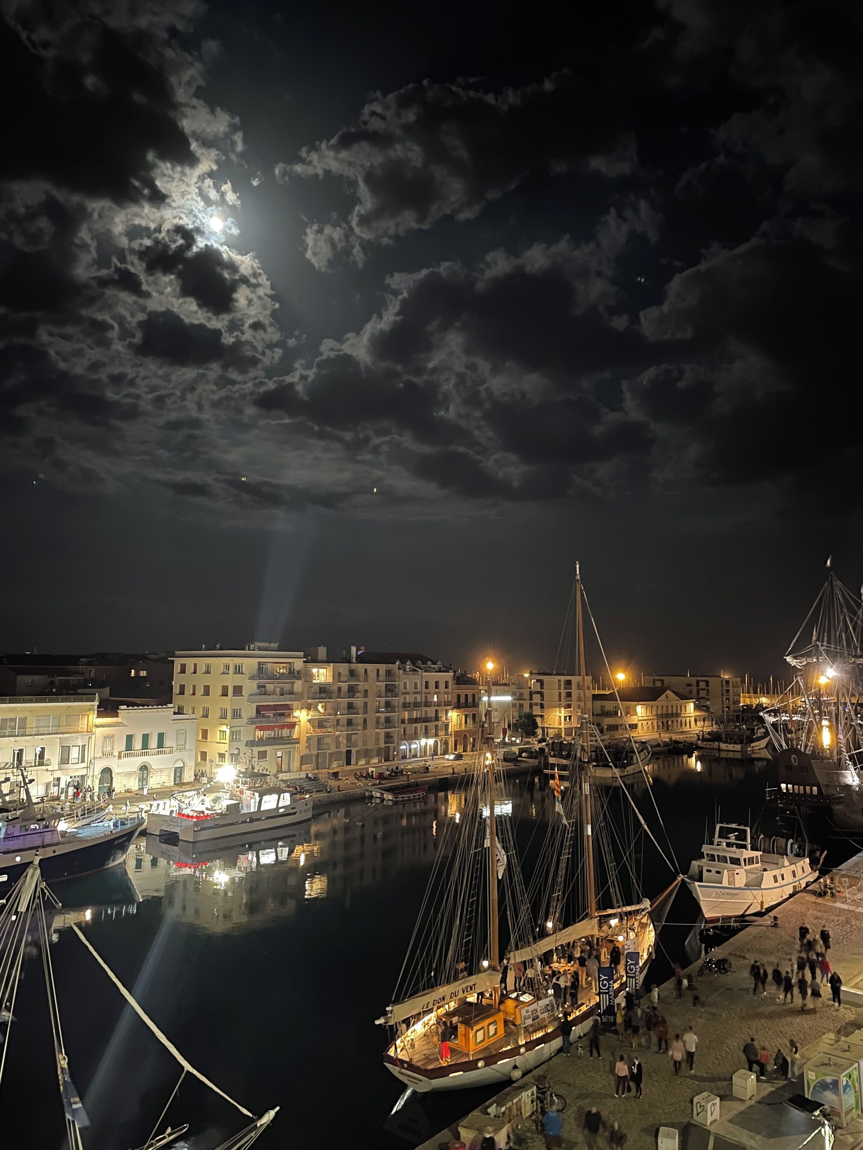 A marina at night with boats docked, a crowd on the pier, and buildings in the background.