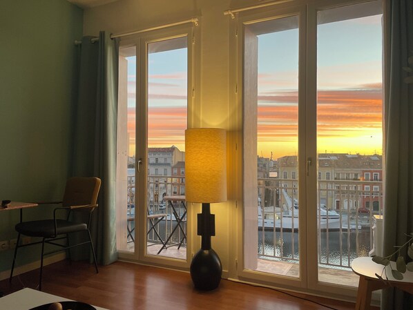 Een kamer met een groot raam, een stoel, een tafel en een lamp.