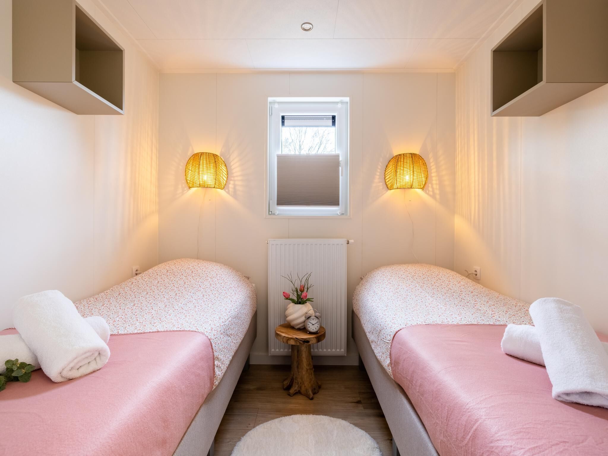 A room with two beds, a small table with a clock, a radiator, and wall-mounted lamps.