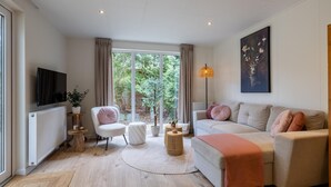 A modern living room with a sofa, a TV, a lamp, and a large window. - Luxury forest stay with covered terrace and BBQ (Hattemerbroek)