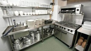 A commercial kitchen with stainless steel appliances, shelves with white dishes, and a sink. - Sky Hotel (Lucknow)