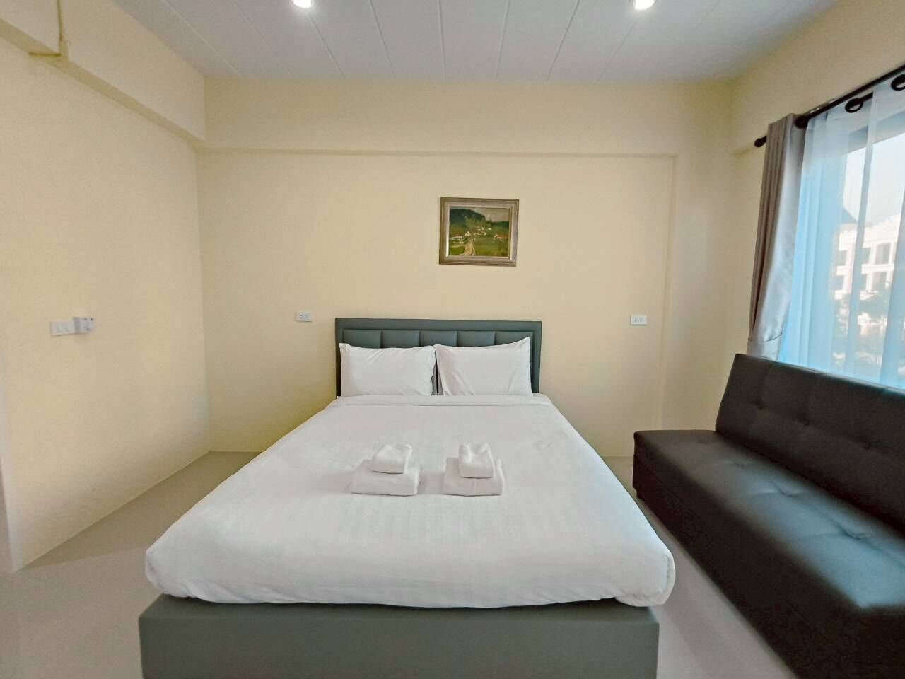 A neatly made bed with white linens and two towels, a dark sofa, and a painting on the wall.
