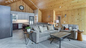 A spacious living area with a sofa, coffee table, and a dining table. A kitchen with a refrigerator and sink is visible in the background. - Sauna & Game Room! Tranquil Retreat on Grace Lake (Watersmeet)