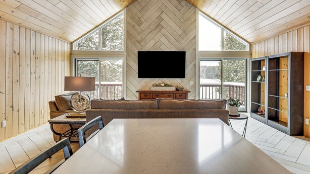 A spacious room with a large dining table, a sofa, a TV mounted on a herringbone-patterned wall, and large windows. - Sauna & Game Room! Tranquil Retreat on Grace Lake (Watersmeet)
