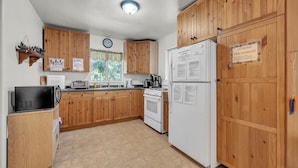 A kitchen with wooden cabinets, a microwave, a stove, and a refrigerator. There is a clock on the wall and a window with a view of trees. - Lakefront Cabin w/ Dock, Kayaks & Full Kitchen (Nevada City)