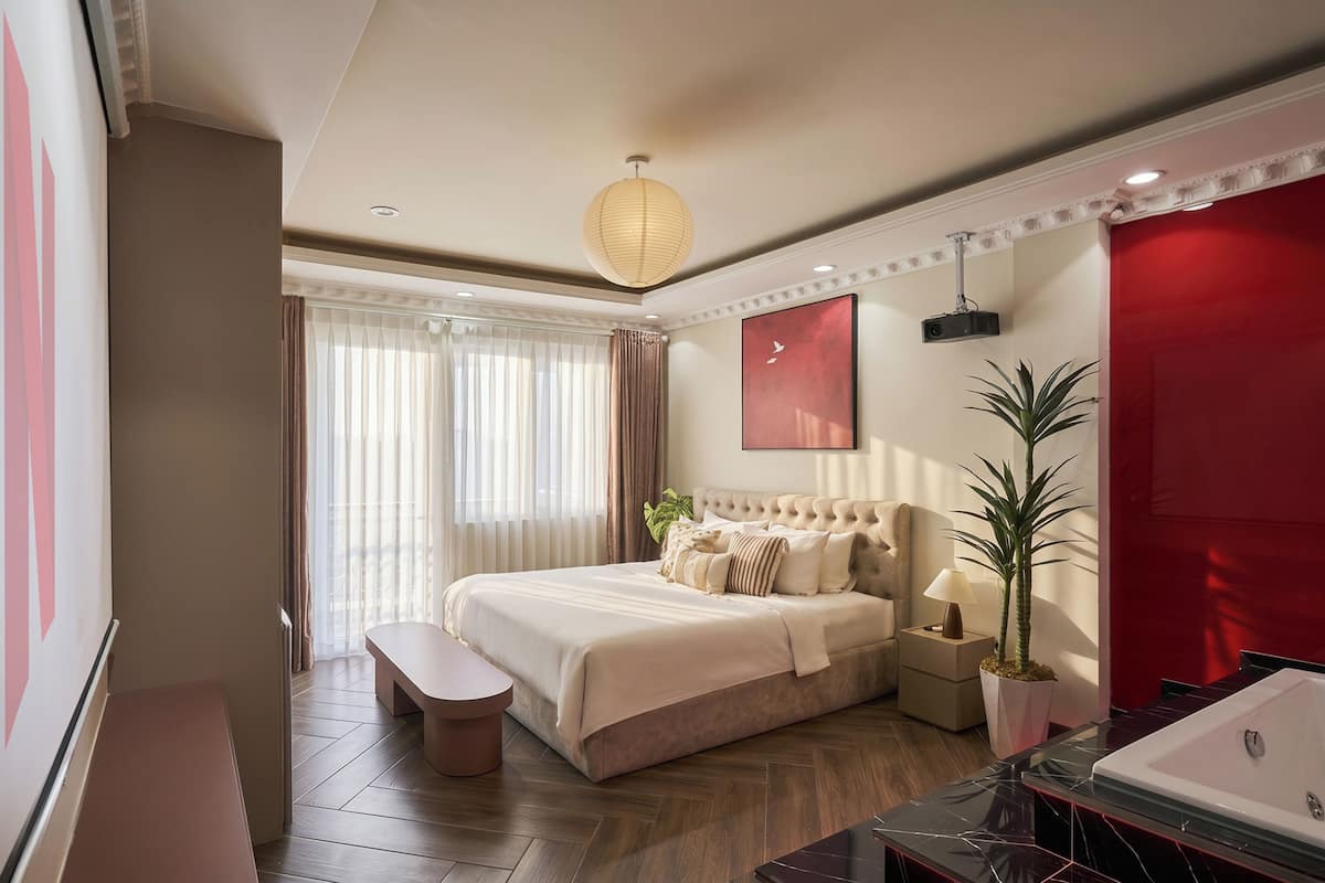 A modern hotel room with a large bed, a bedside table with a lamp, a red accent wall, and a large window with curtains.