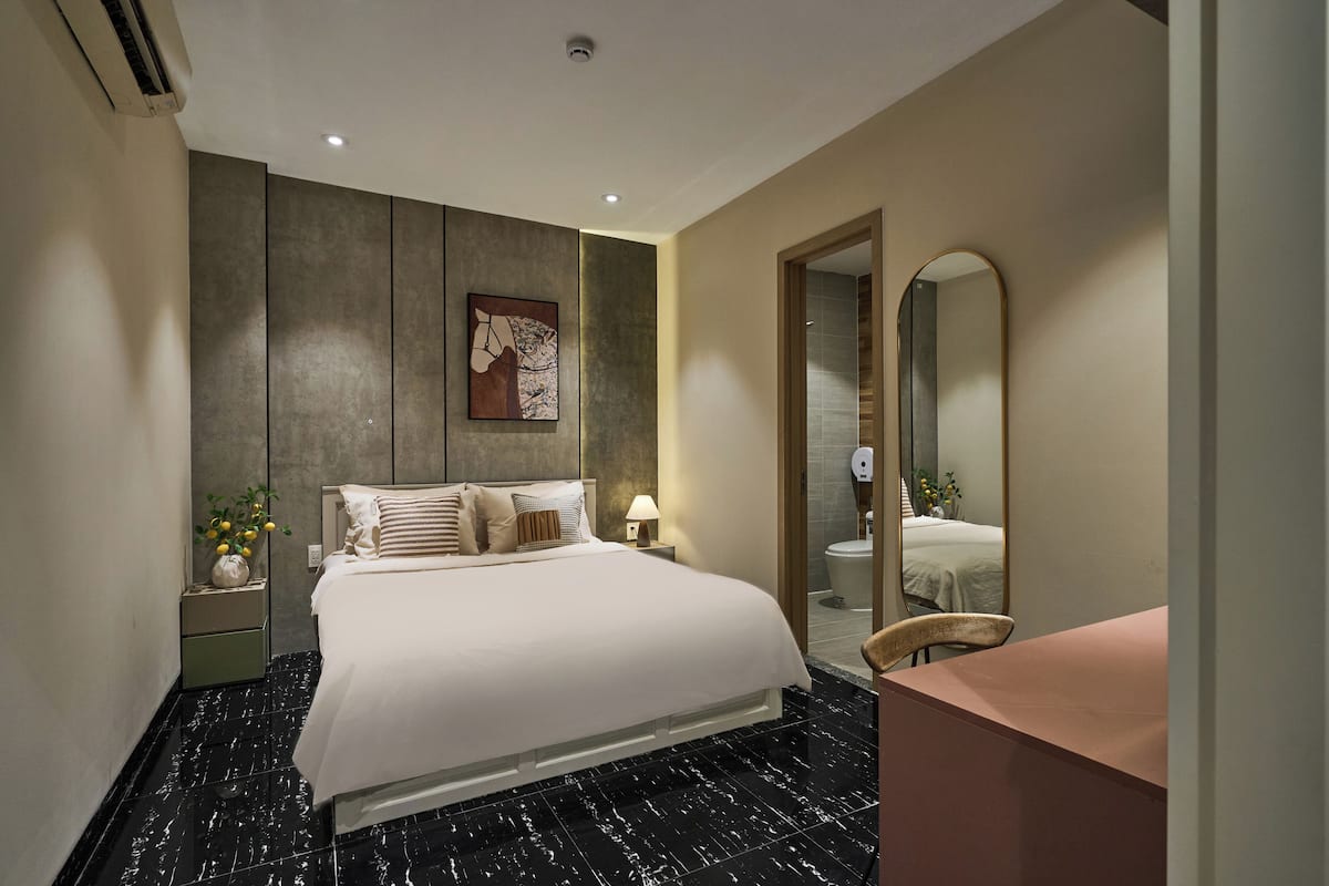 A modern hotel room with a large bed, a bedside table with a lamp, a mirror, and a small seating area.