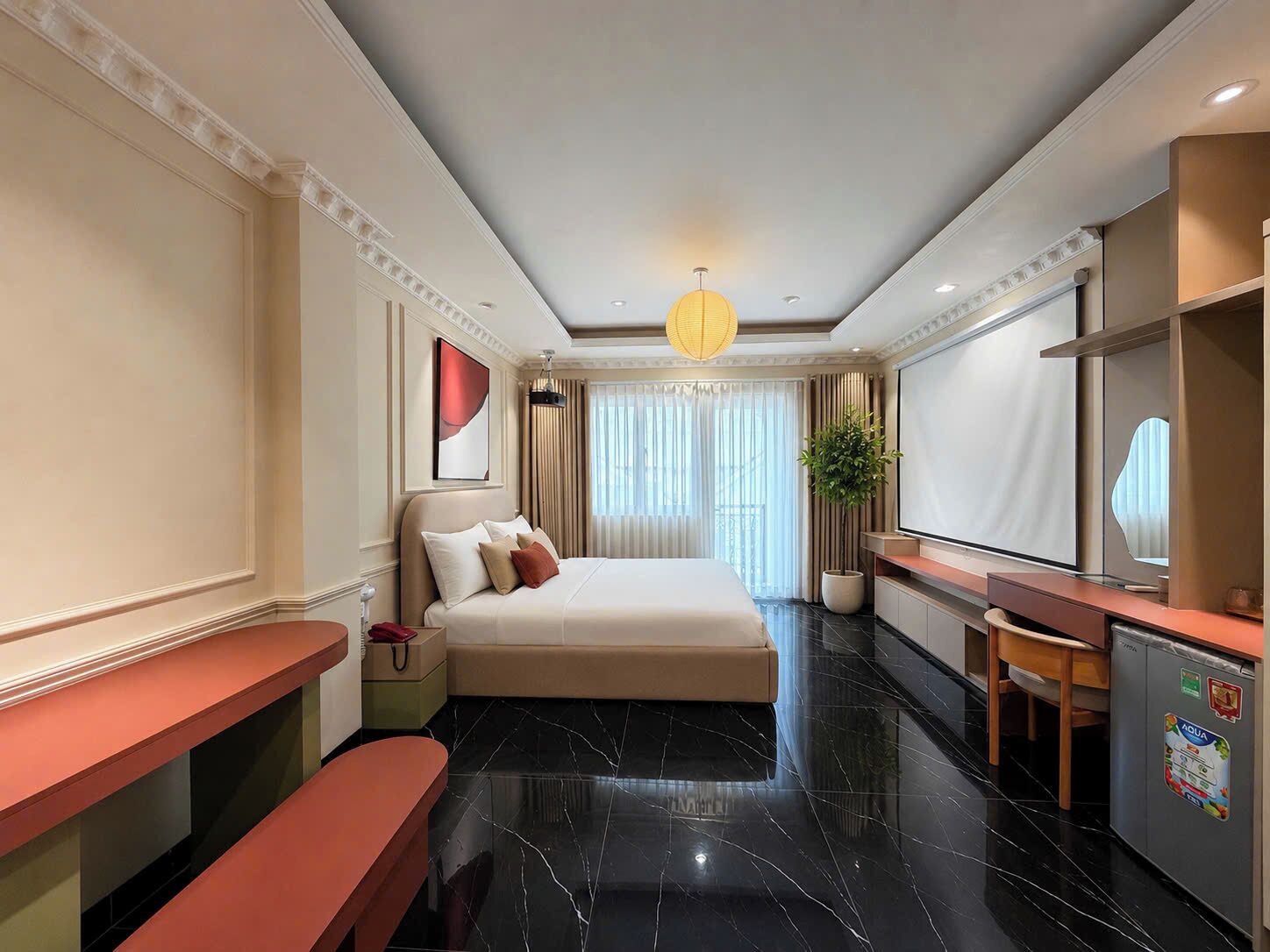 A hotel room with a bed, a desk, a bench, a large mirror, and a potted plant.