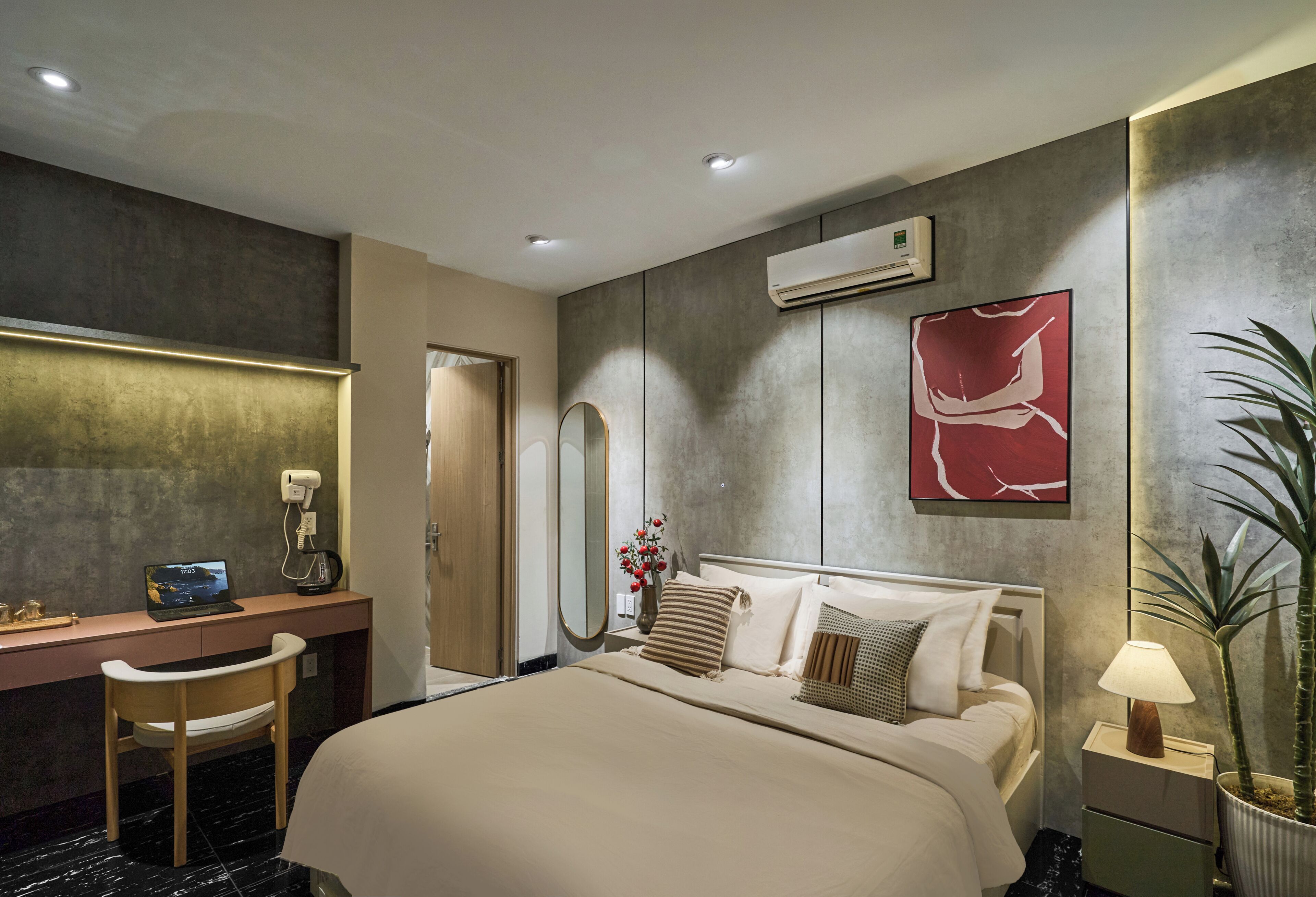A hotel room with a bed, desk, chair, bedside table, lamp, potted plant, and artwork.