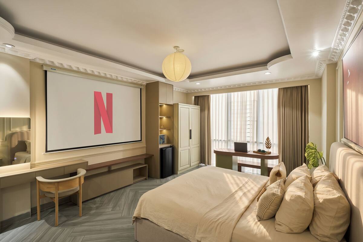 A hotel room with a large bed, a desk with a chair, a TV with a Netflix logo, a plant, and a window with curtains.