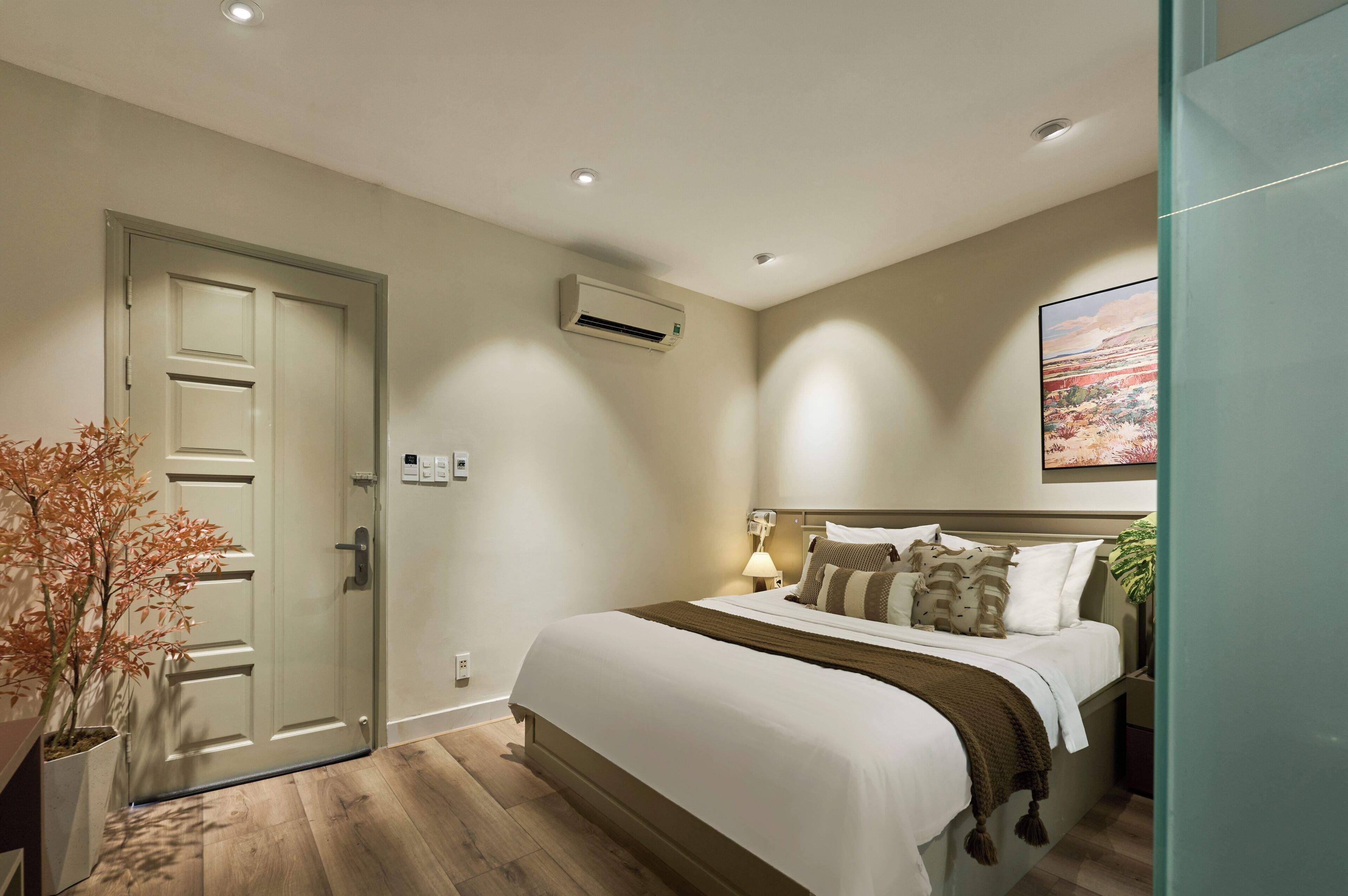A hotel room with a bed, bedside lamp, air conditioning unit, and a painting on the wall.