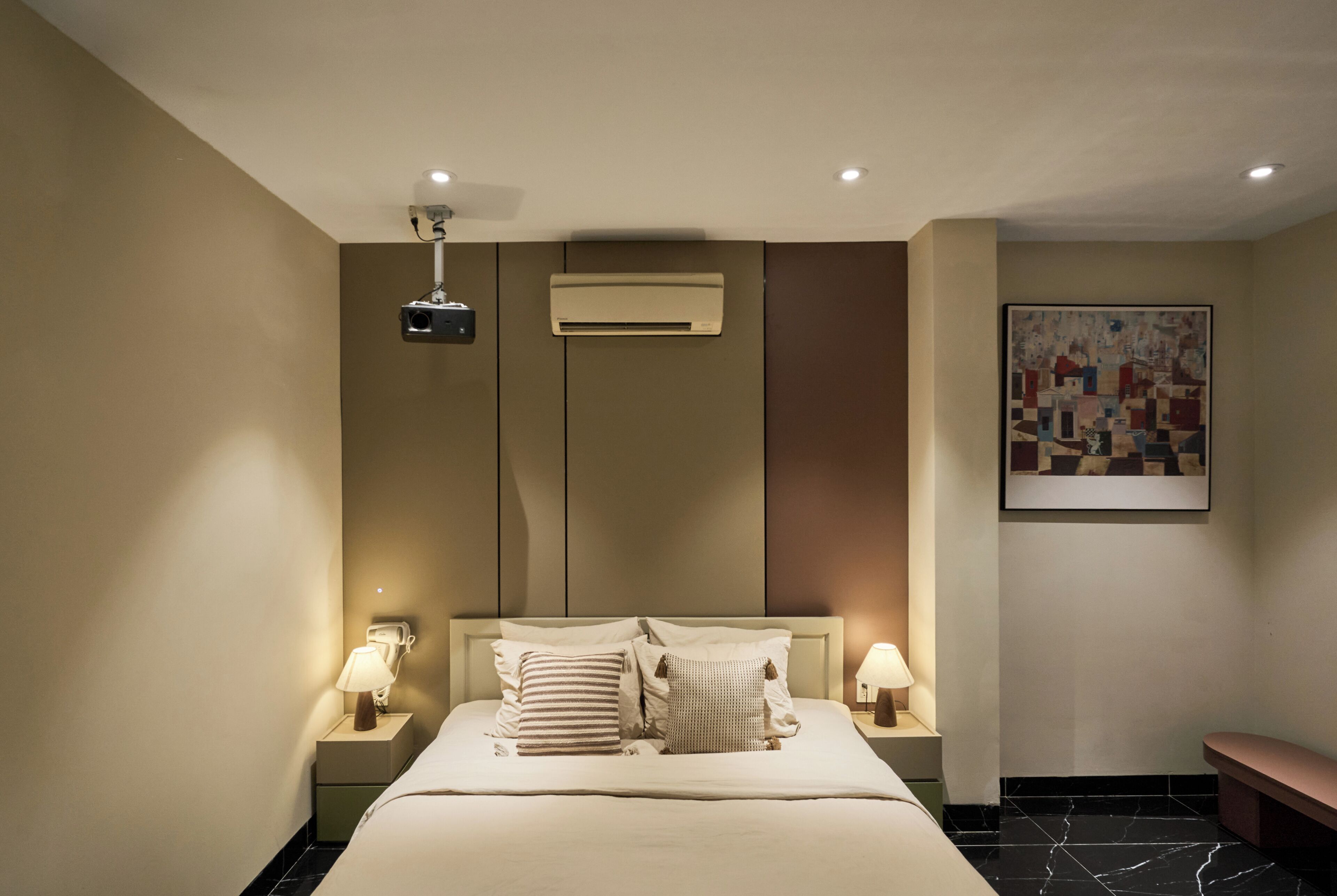 Une chambre d’hôtel équipée d’un lit, de deux lampes de chevet, d’un climatiseur, d’un projecteur fixé au plafond et d’un tableau abstrait encadré accroché au mur.