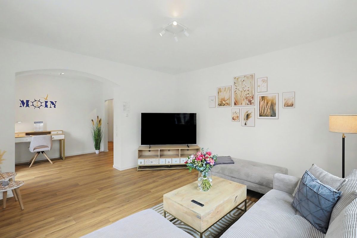 A modern living room with a flat-screen TV, a sofa, a coffee table with a vase of flowers, and a desk with a computer.