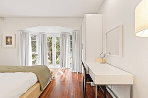 Ein Schlafzimmer mit Bett, Schreibtisch mit Spiegel und Blick ins Freie durch ein großes Fenster mit Vorhängen.