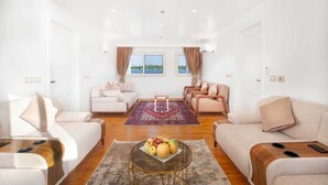 Een ruime woonkamer met een groot raam, een houten salontafel met fruit, en een rood, geruit tapijt.