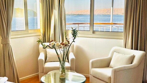 Hotelkamer met een groot raam met uitzicht op zee, twee witte fauteuils, een glazen salontafel met een bloevas en een bed met witte lakens.