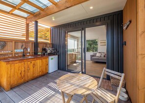 Una terraza de madera con barra integrada, una mesa redonda y un ventanal corredero que da a un espacio de estar al aire libre.