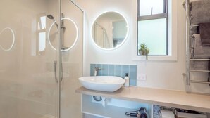 Une salle de bain moderne avec une douche à parois de verre, un lavabo blanc et un meuble-lavabo doté d’un miroir et d’une fenêtre au-dessus.