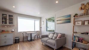 Un salon moderne avec un canapé gris, un plancher en bois, une table à manger avec des chaises, une bibliothèque et une grande fenêtre laissant entrer la lumière naturelle.