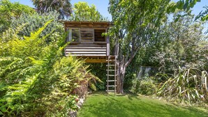 A wooden treehouse with a ladder, surrounded by lush greenery and a well-maintained lawn.